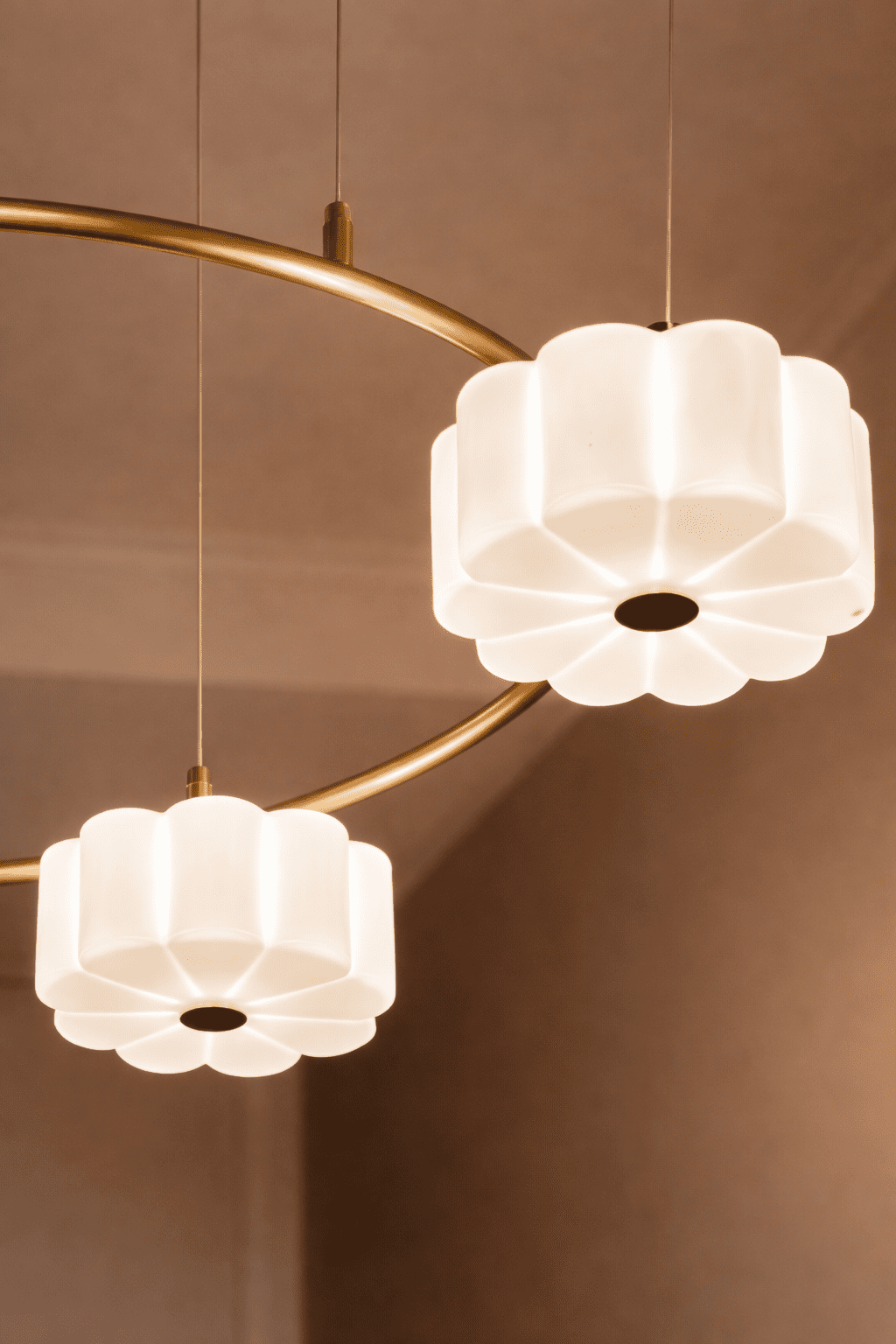 Détail vertical d’une suspension Art Déco en laiton brossé, montrant deux opalines en forme de fleurs, suspendus par de fins câbles métalliques, avec une lumière douce et chaleureuse et un arrière-plan subtilement flouté.